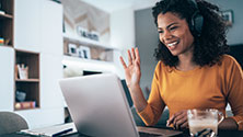 Woman wearing headphones smiling and waving at a laptop screen.