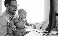 Man holding a baby while smiling at a computer screeen.