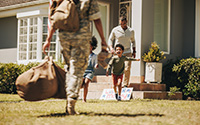 Veteran carrying equipment walking across a lawn to their house where a family is rushing out to greet them.